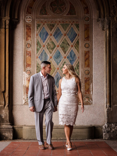 Engaged couple walks hand-in-hand beneath the Bethesda Terrace arcade in Central Park, NYC, captured in a romantic moment by NYC wedding photographer Heritage House.