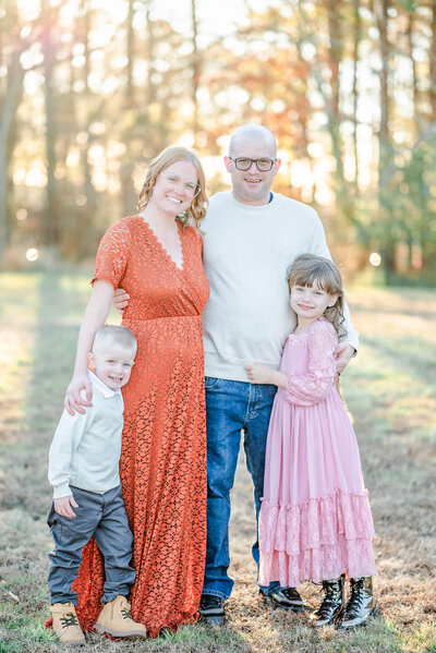 A family puts their arms around each other and smile for the camera. The mom is in an orange lace dress. The daughter wears a long pink dress. The dad and son are both in jeans with white sweaters.
