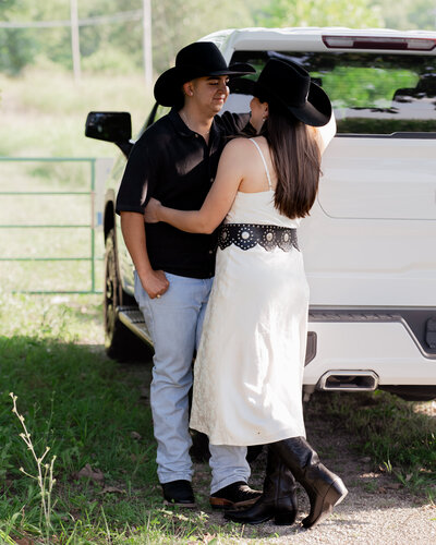 a southern catholic couple embrace during their engagement session while wearing cowboy hats and standing next to a truck, showing the elegance of the photography style contrasted with the fun and relaxed nature of southern couples