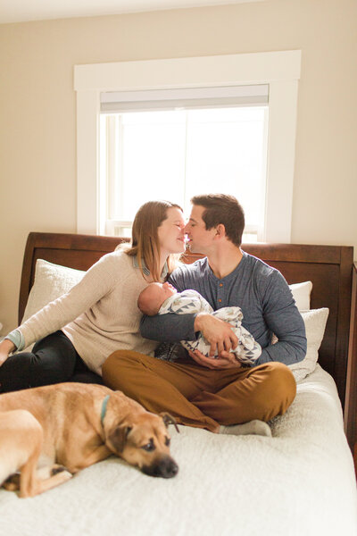 man and woman holding newborn baby sitting near dog in bed kissing
