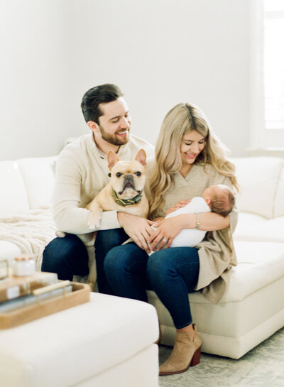 Family with their dog sits on their white couch for a photo during their downtown Raleigh newborn session. Photographed by Raleigh newborn photographers A.J. Dunlap Photography.