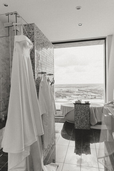 Wedding dress hanging in a modern hotel bathroom at the Westin in downtown Dallas. The space features sleek tile, a city-view window, and a freestanding tub, capturing a quiet moment before the day begins.