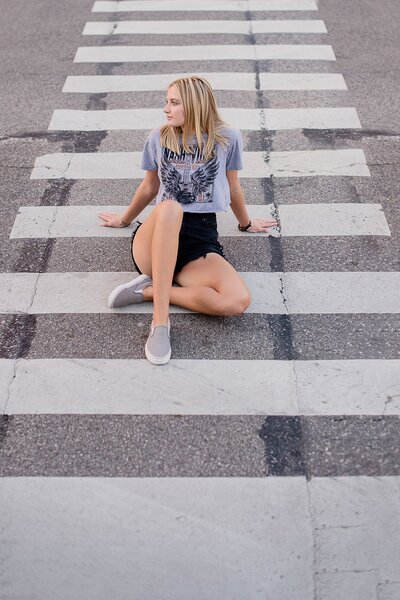teenage blonde girl in gray t-shirt and black jean shorts sitting in street cross walk