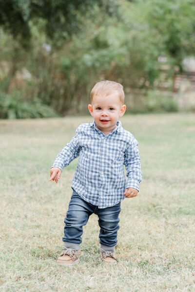 A little boy dressed in a blue plaid shirt and blue jeans walk on grass at Katherine Fleischer Park. He looks at the camera with a smile on his face in this candid shot taken by a photographer specializing in family portraits in Austin, TX