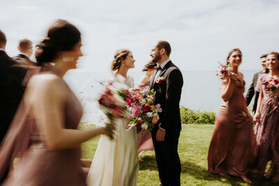 A photo of a wedding couple at their outdoor weddings looking towards each other standing close holding flowers with their wedding party surrounding them