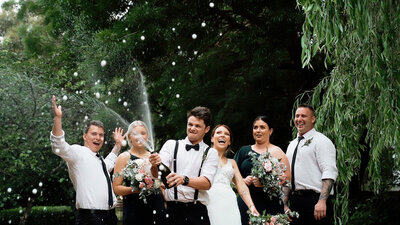 groom opens and sprays Champaign on wedding day surrounded by bride and wedding party