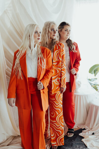 Kenzi in an orange suit, Sophie in a multicolored orange suit, and Marisa in a red suit standing and looking over their shoulders