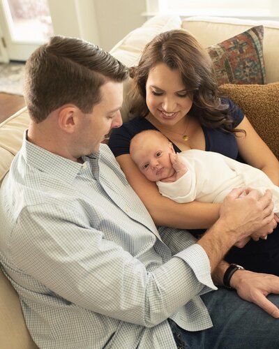 Loving parents gaze at their smiling newborn, swaddled in white, during an in-home newborn session with Moments in Light Photography.