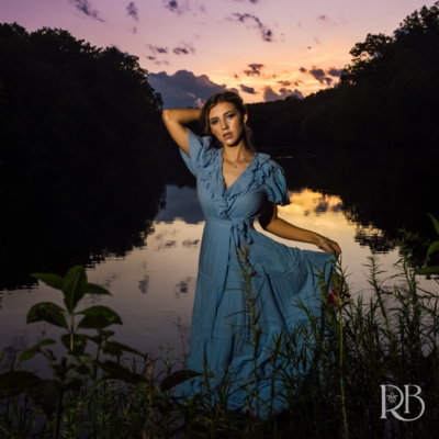 girl in a long blue dress standing in a lake at sunset for her high school senior portrait; the sunset is purple and pink and there are trees and hills behind her in shadow