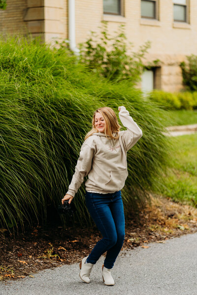 Staci striking a playful pose in a hoodie and jeans surrounded by tall greenery, showing off her fun and adventurous personality.
