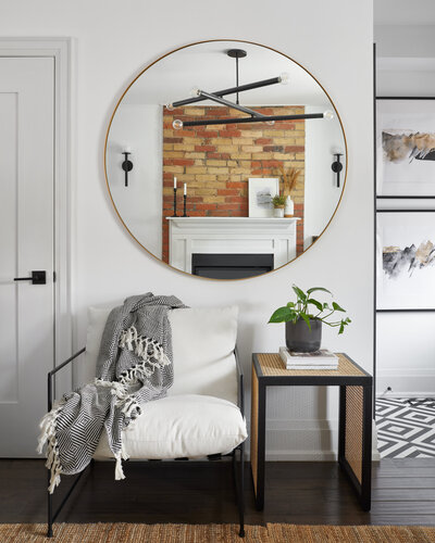 A black and white patterned throw blanket on top of a plush white chair in a bright room with white walls and dark brown hardwood flooring. A rattan side table with a potted plant sits next to the chair and a large circular mirror hangs on the wall above.