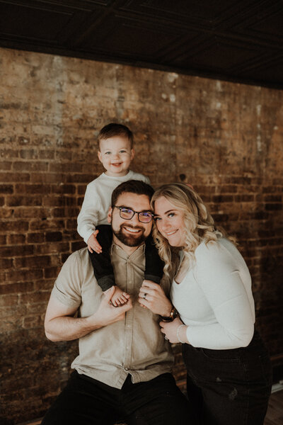 Family photoshoot of Morgan Ashley Lynn Photography owner, Morgan; her son is sitting on her husband's shoulders smiling and she is hugging her husband's arm with their heads together standing in front of a brick wall for a vintage look