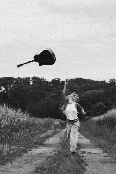 A person in a hat, shirt, and boots throws a guitar in the air while walking on rocky desert ground with mountains and sparse vegetation in the background—captured in black and white for Sweet Pea Ink Creative’s web design inspiration.