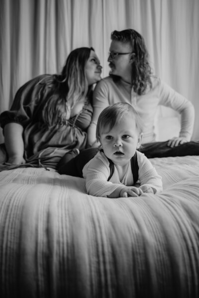 Black and white photo of baby laying on bed in front of mom and dad who are behind him looking at each other in Annapolis Maryland.