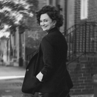 A black and white portrait of a woman with short, curly hair, looking back over her shoulder with a slight smile. She is walking on a city street, and the background is blurred, giving a sense of motion.