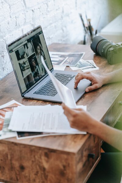 A person working at a wooden desk, reviewing printed documents while using a laptop. The laptop screen displays several images, and a camera with a large lens is placed next to the laptop. The workspace is bright, with a rustic, creative feel.