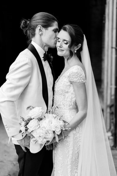 Black and white portrait of a groom kissing his bride's forehead while she holds a bouquet, both looking joyful.