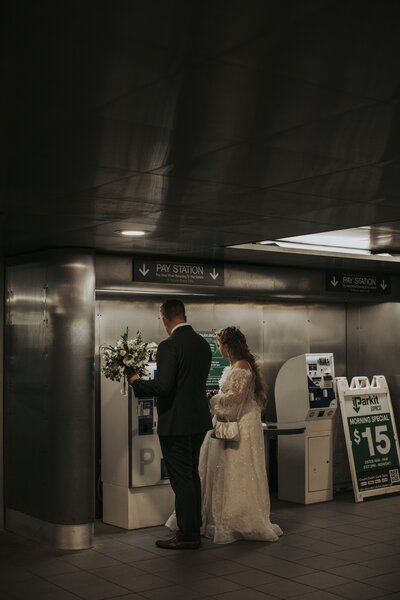 A bride and groom in wedding attire stand at a parking payment kiosk in a dimly lit garage, holding a bouquet while handling the ticket machine together.