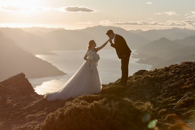 Bride and groom on top of a mountain in New Zealand with a large lake and other mountains in the distance. The groom is kissing the hand of his bride.