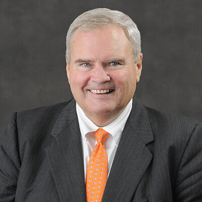 Smiling senior man in a dark gray pinstripe suit, white shirt, and bright orange patterned tie, posed against a plain dark background in a professional headshot.