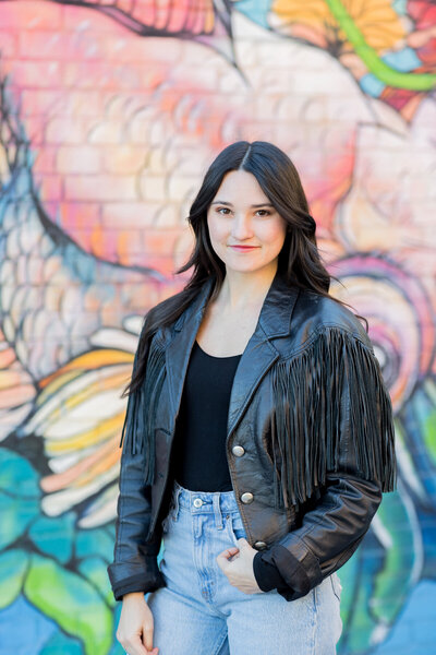 Confident woman in a black leather fringe jacket and light-wash jeans poses in front of a vibrant, artistic mural in Columbia, SC. Her modern headshot showcases a stylish, urban look, perfect for personal branding and professional portraits.