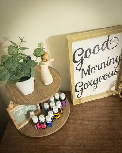 Essential oils and greenery on wooden shelf with a framed "Good Morning Gorgeous" sign in a cozy wellness workspace.