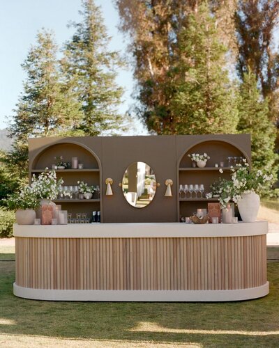 Outdoor bar with semi-circular wooden facade, arched shelves, a central oval mirror, and decorative vases with white flowers in a grassy area.