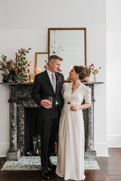 A bride and groom in a white wedding gown and Black suit staring into each other’s eyes in front of a fireplace.