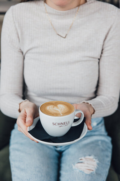 Mitarbeiterin gießt Milch in einen Cappucchino und macht mit Latte Art ein Herz - Café Schnell