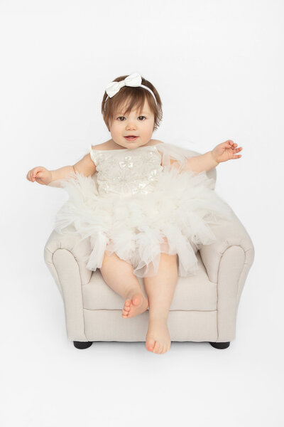 studio one year portrait of a happy one year old in a sequined and tulle dress, with bare feet; she is pictured in a cream colored leather chair for children
