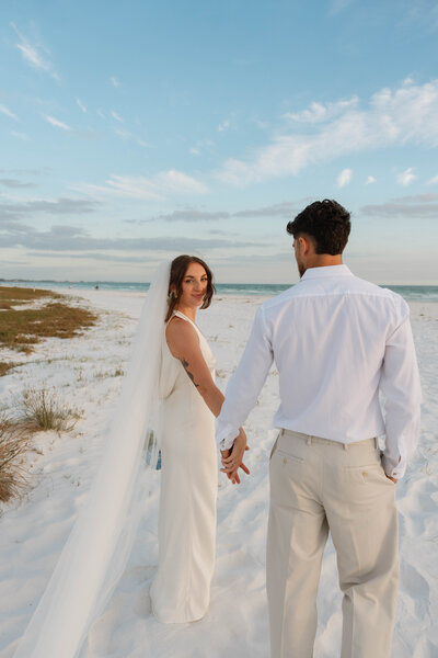 a bride and groom walking on a white sandy beach 