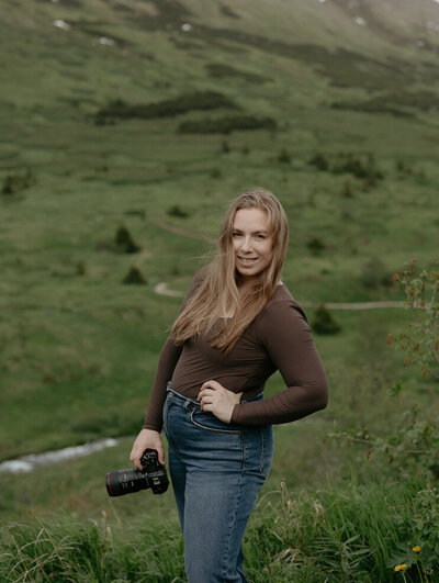 Woman holding a camera and wearing a brown shirt and blue jeans.