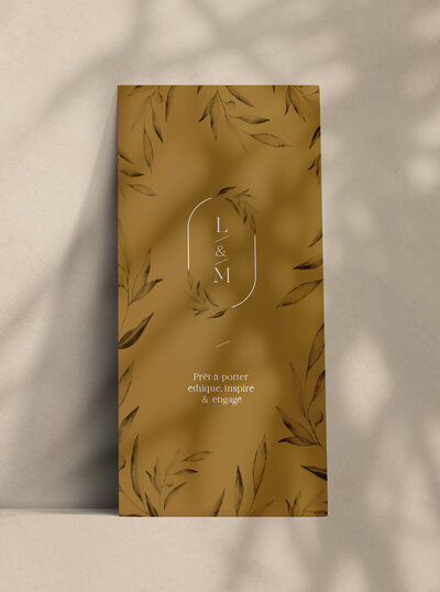 Carton d'invitation ocre, format vertical, reposant sur un mur avec illustration de feuilles en aquarelle. Support imprimé crée par le Studio graphique en Vendée.