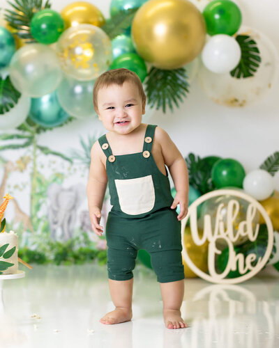A happy baby boy is dressed all in green standing in front of a jungle cake smash set, smiling and looking at the camera
