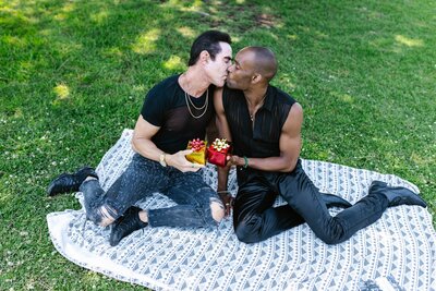 Two masculine presenting partners sit on a picnic blanket, surrounded by green grass in the background. They learn across the blanket to kiss each other, both holding smalls gifts in their hands - one with yellow wrapping paper, another with red wrapping paper.