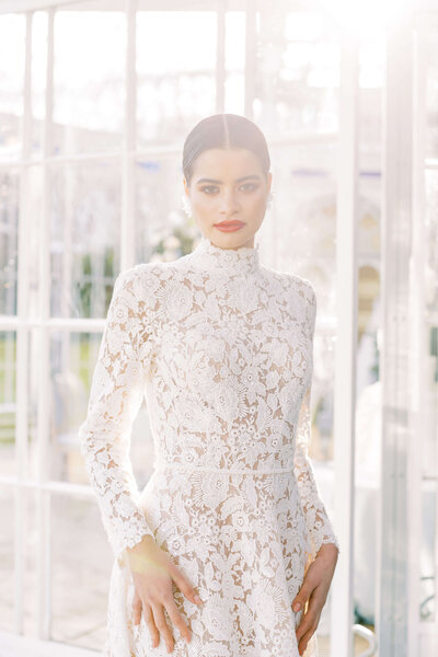 bride poses in soft lighting wearing a lace dress and red lipstick outside a luxury wedding venue glasshouse