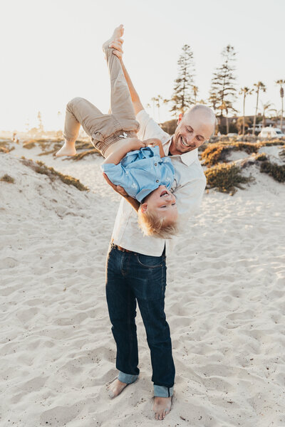 a dad holds his son upside while they both laugh on the beach during their family photos in Coronado