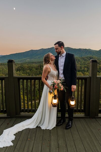 A bride and groom stand side by side with their arms around each other. They are looking lovingly at each other after eloping. They are standing in front of a vast mountain range as the sun sets painting the sky pink, blue and orange as the moon rises behind them.