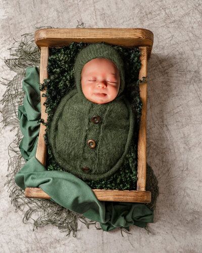Baby boy wrapped in a green swaddle with brown buttons sticks his tongue out at the camera