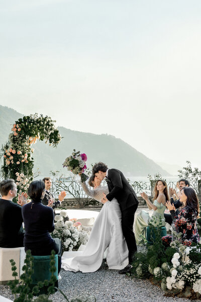 Bride and groom sharing first kiss under floral arch during intimate Lake Como wedding ceremony with guests seated along the waterfront at Villa Pizzo