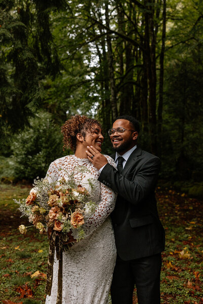 Groom smiling and touching bride’s face as she holds a romantic bouquet of orange and white flowers during their woodland elopement in Oregon.