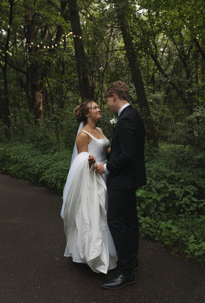 Bride and groom getting portraits on a path in the middle of the trees, looking at each other with love in their eyes. There are lights above them lighting them up as they prepare to walk down the isle