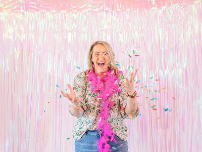 A person wearing a floral shirt and a purple feather boa is joyfully throwing colorful confetti in front of a shimmering pink backdrop, celebrating their VIP launch day for an exquisite Showit website template for women entrepreneurs.