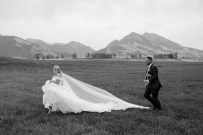 Black and white photo of bride and groom running through a Montana field, her dress and veil flowing in the wind.