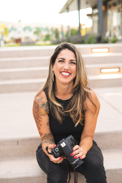 Vanessa Vancour, a Latinx woman with long hair, sitting outside, holding a camera and smiling.