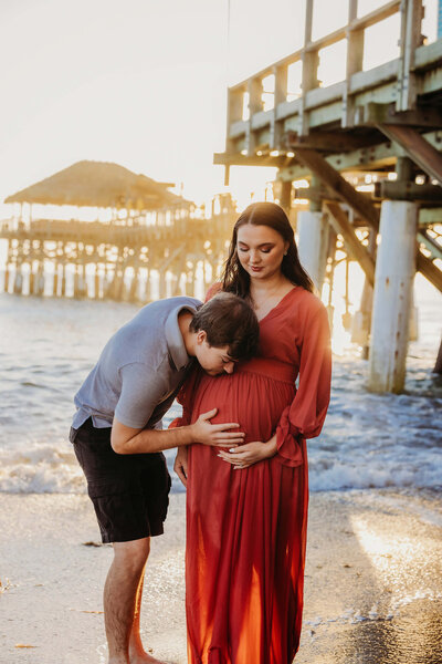 A man gently kisses a pregnant woman's belly in Cocoa Beach at sunrise. She is wearing a long red dress, and they stand near a wooden pier, with gentle waves lapping at the shore—perfect for a beach maternity photographer to capture this serene moment.