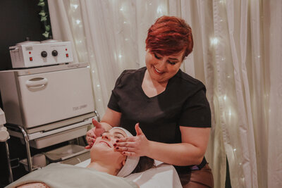 Brittney is giving her client a facial with her hands and smiling. Her client is also smiling while getting the facial and looks pleased