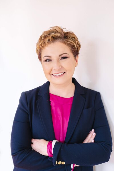 Portrait of a smiling photographer with short hair, wearing a navy blazer over a bright pink top, standing confidently with arms crossed