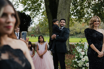 A wedding guest holds a child while standing beside flower girls in pink dresses, with other guests watching the ceremony in a green, tree-filled outdoor space.