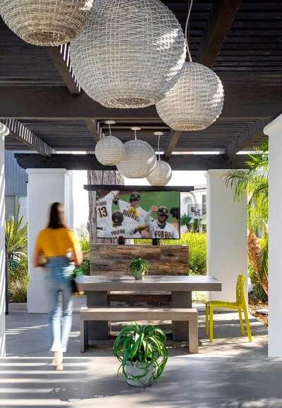 Outdoor entertainment area featuring woven light fixtures, a built-in TV, and modern picnic-style seating surrounded by greenery.